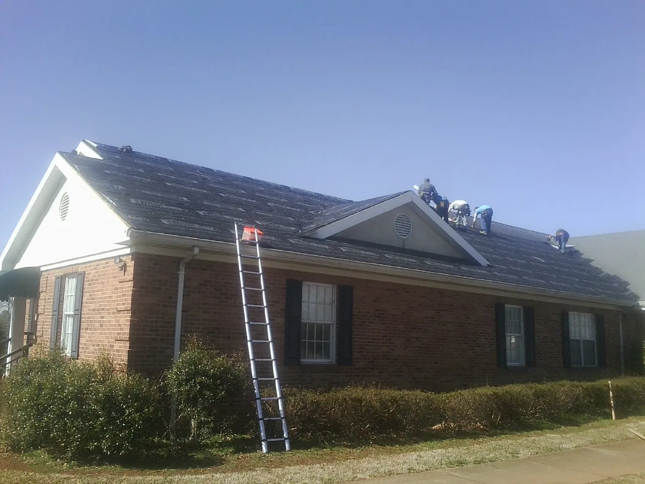 Crew tearing shingles off a brick ranch roof with a ladder set — Metal Roof Repair work by our crew in Cherry Plain