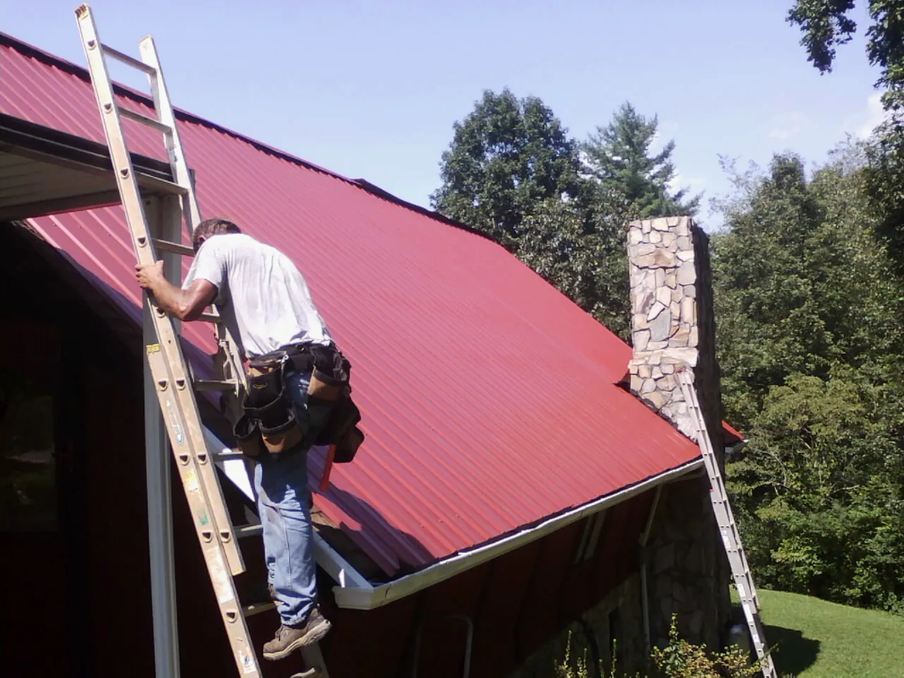 Roofer on a ladder beside a red metal roof and stone chimney — Soffit & Fascia Repair by Roofing Brothers in Cherry Plain